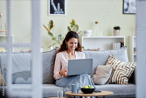 Front view of smiling young woman using laptop while sitting on sofa at home and using internet or working online