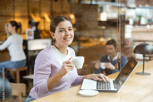 Smiling young woman using laptop and drinking coffee in modern cafe
