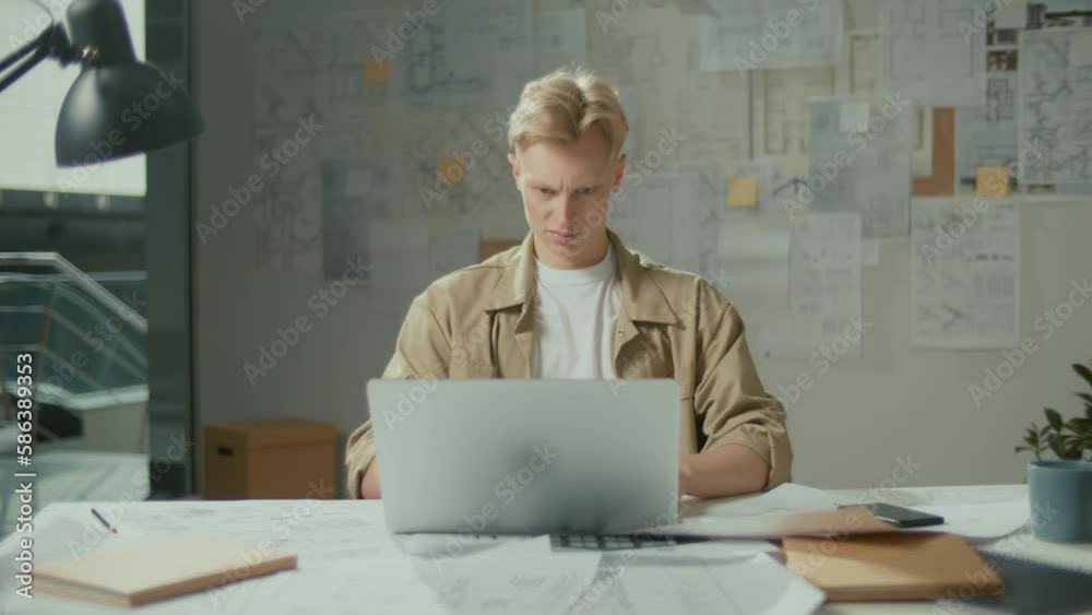 Young Confident Manager Architect Typing on Laptop Working on the Project, Smiling at the Screen and Making Winning Gesture.Handsome Man Architect Doing Project Sitting at Bright Workplace and Smiling Young Confident Manager Architect Typing on Laptop Working on the Project, Smiling at the Screen and Making Winning Gesture.Handsome Man Architect Doing Project Sitting at Bright Workplace and Smiling