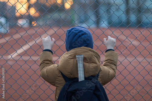 the boy looks at the tennis court holding the links of the fence. rear view image