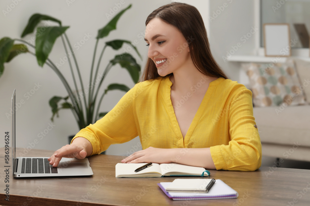 Happy young woman with notebook working on laptop at wooden table indoors Happy young woman with notebook working on laptop at wooden table indoors