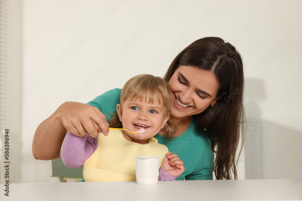 Mother feeding her cute little child with yogurt at white table in room