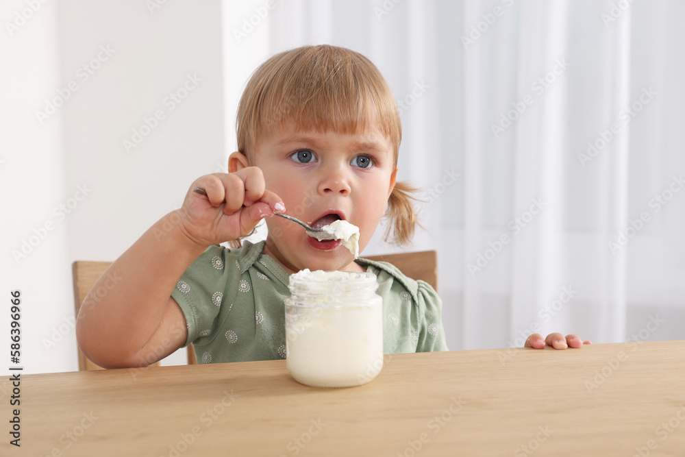 Cute little child eating tasty yogurt with spoon at wooden table indoors Cute little child eating tasty yogurt with spoon at wooden table indoors