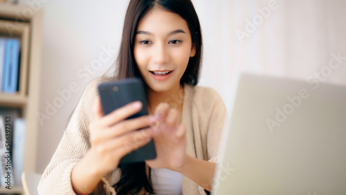 Young asian woman happy working on computer laptop and hand typing chat message on mobile smartphone at home. Video call  Online meeting