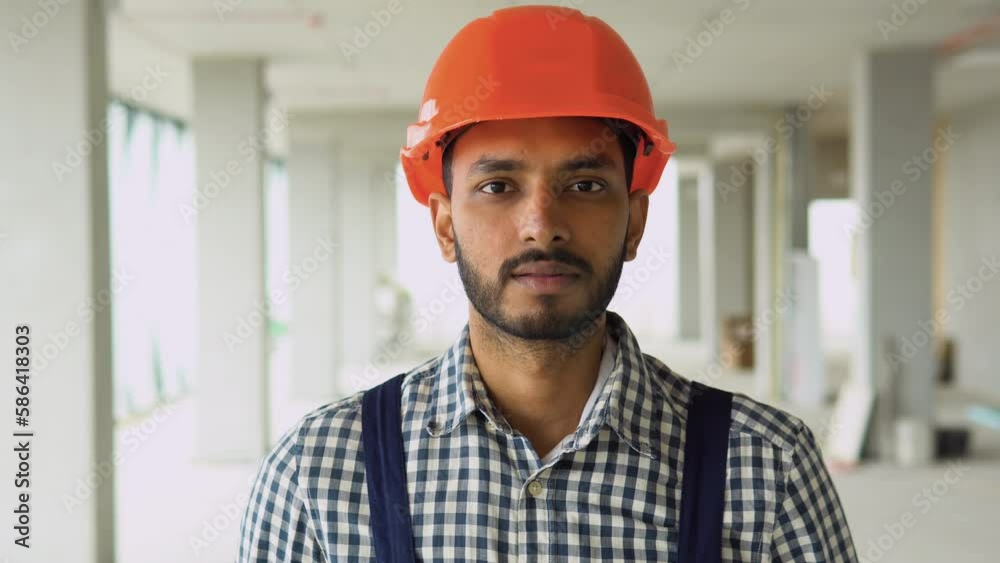 Portrait of Indian or Pakistani worker builder in uniform wearing a safety helmet looking at the camera, standing on construction site Portrait of Indian or Pakistani worker builder in uniform wearing a safety helmet looking at the camera, standing on construction site