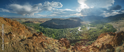 View of a green mountain valley among dry slopes, meanders of the river, against the sun