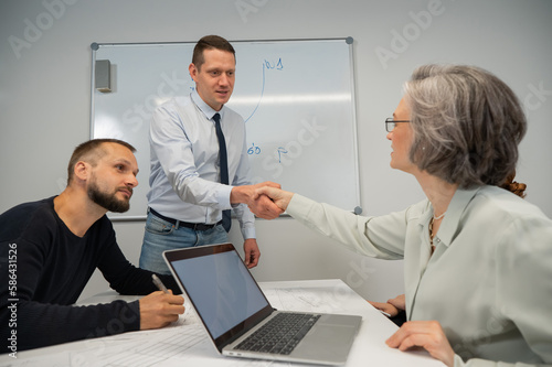 The boss makes a presentation to subordinates at the white board. Caucasian man shaking hands with middle aged woman. 