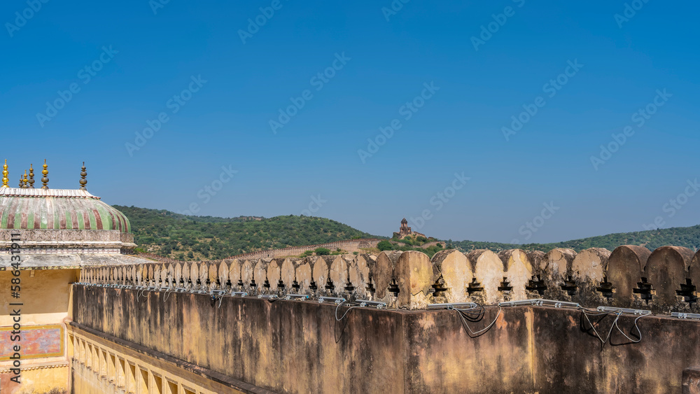 The upper part of the weathered wall of the ancient Amber Fort. A dome with spires on a blue sky background. In the distance, on the crest of the mountain-a fortress wall with a watchtower. India.