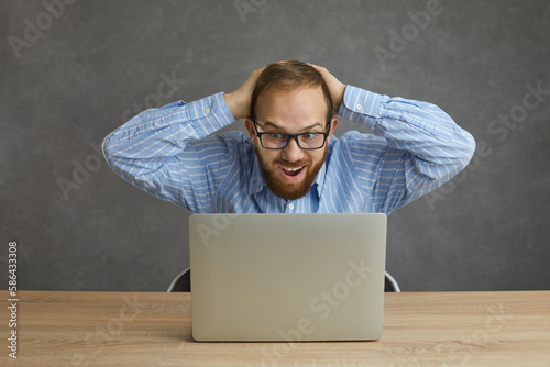 Happy shocked caucasian man with impressed face expression screaming touching head with hands looking at laptop screen. Surprised stock broker, freelancer entrepreneur work online. Studio portrait