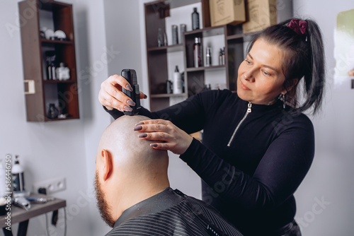 Girl hairdresser shaves an adult man with beard baldly using electric razor in barbershop holding it in her hand. Provision of hairdressing services. photo