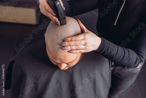 Photo from top to bottom. Man with beard sits in chair with cape in barbershop to get rid of hair from his head completely.