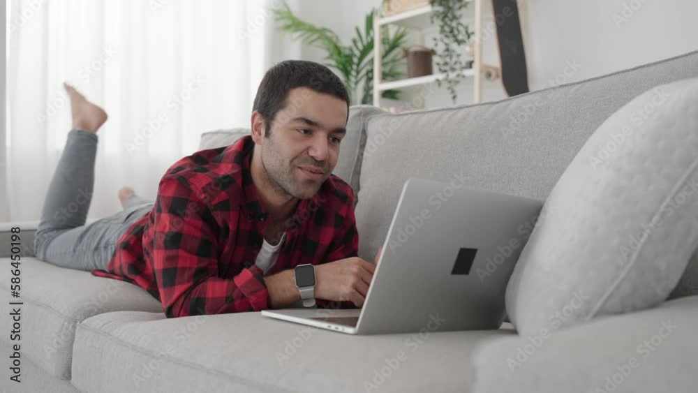 Hispanic young man having video call on laptop lying down on sofa. Hispanic young man having video call on laptop lying down on sofa.