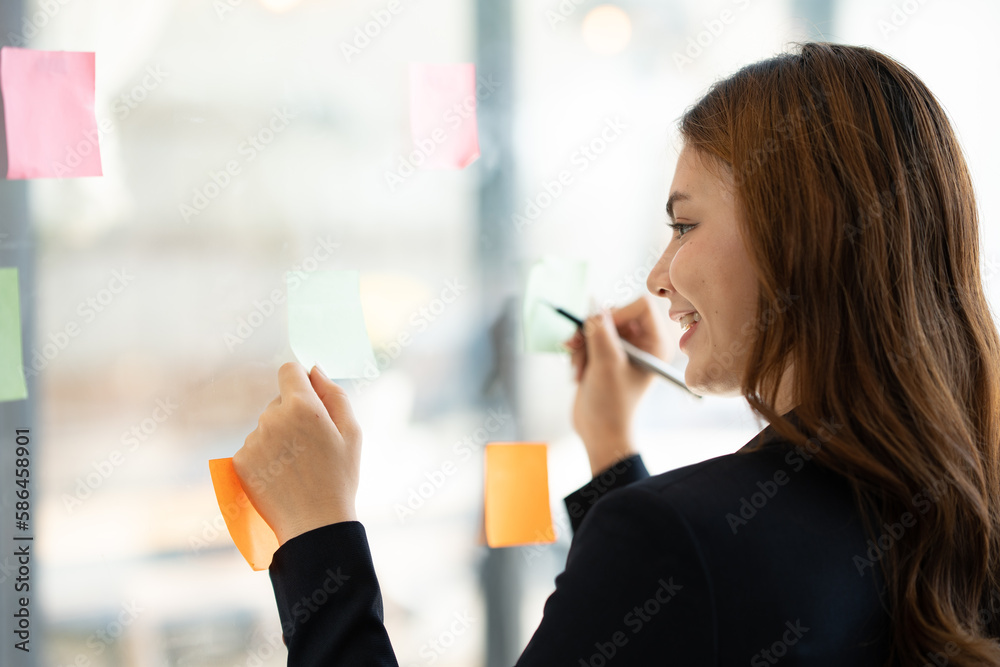 Happy asian businesswoman working with sticky notes on glass wall as reminder and record work orders in financial business online job tax Plan a meeting in the office. Happy asian businesswoman working with sticky notes on glass wall as reminder and record work orders in financial business online job tax Plan a meeting in the office.