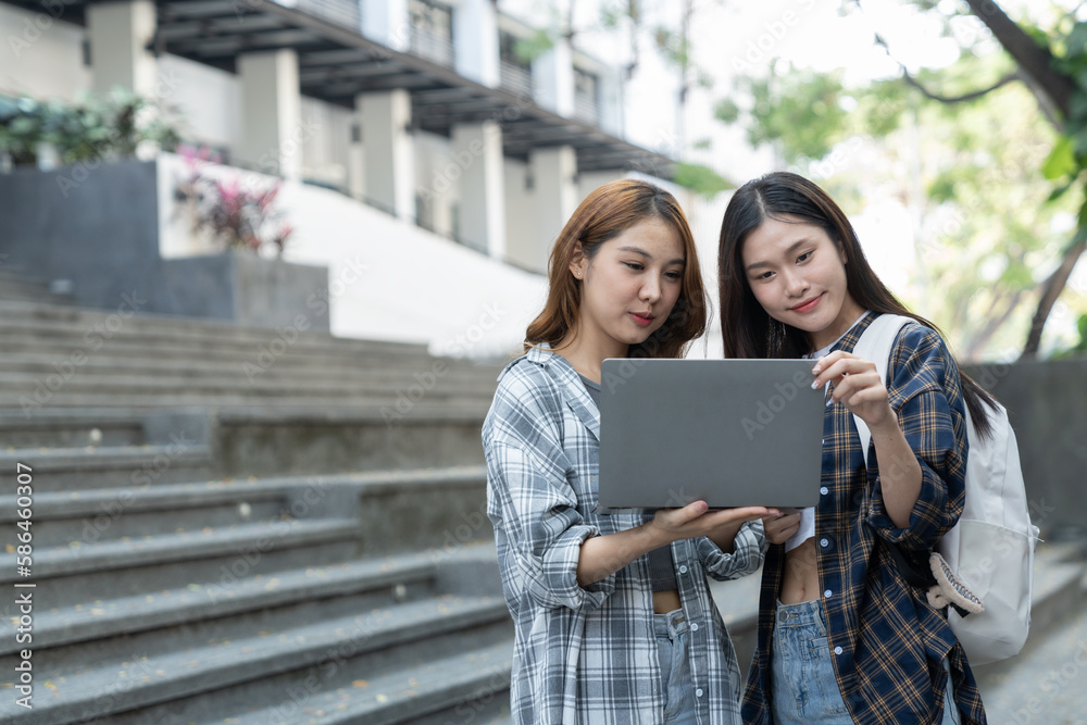 Fototapeta premium Asian female college students look at and study insights and biology on a laptop and record in a notebook at the outside university.