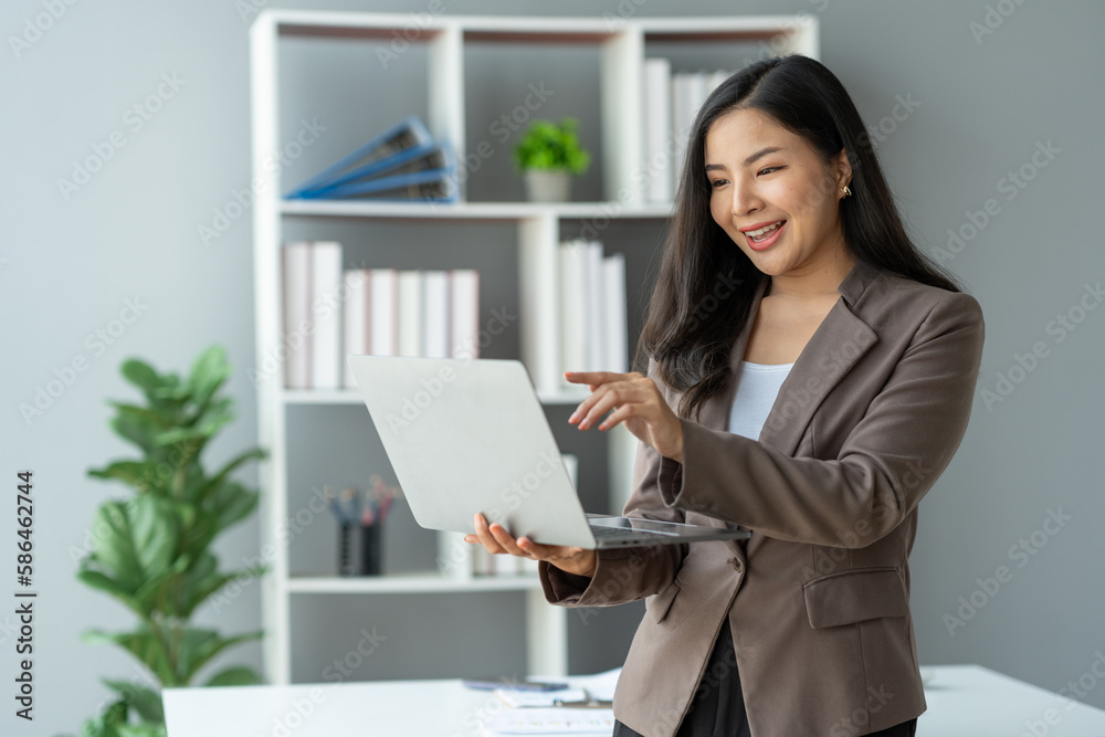 Asian businesswoman smiling confidently successful entrepreneur in the financial business professional company executive Wearing suit standing holding laptop searching business information in office Asian businesswoman smiling confidently successful entrepreneur in the financial business professional company executive Wearing suit standing holding laptop searching business information in office
