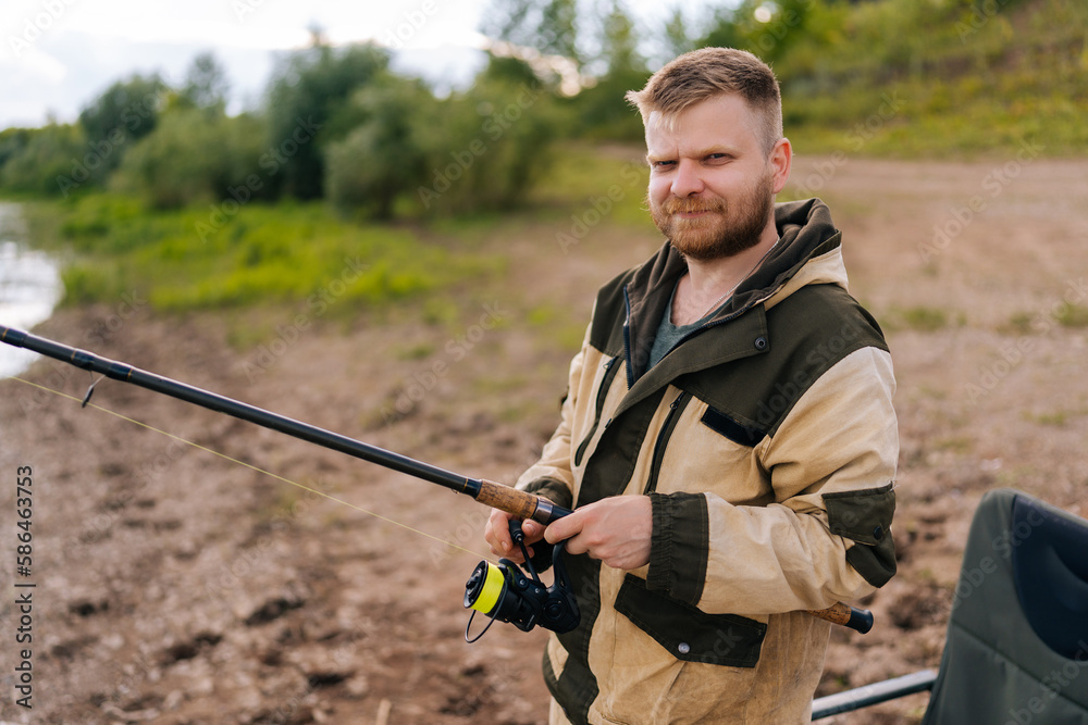 Portrait of smiling fisher man holding spinning casting rod standing on bank waiting for bites on water river at summer day, looking at camera. Concept of lifestyle, leisure activity on nature Portrait of smiling fisher man holding spinning casting rod standing on bank waiting for bites on water river at summer day, looking at camera. Concept of lifestyle, leisure activity on nature