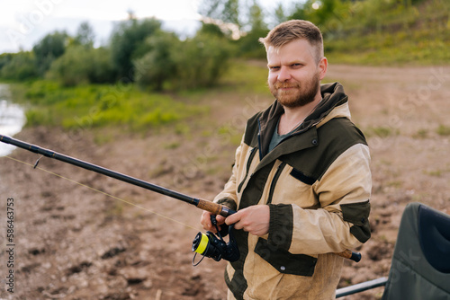 Portrait of smiling fisher man holding spinning casting rod standing on bank waiting for bites on water river at summer day, looking at camera. Concept of lifestyle, leisure activity on nature