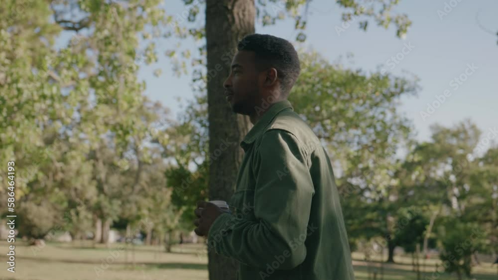 Young black man in shirt walking with coffee in disposable cup under trees in park Young black man in shirt walking with coffee in disposable cup under trees in park