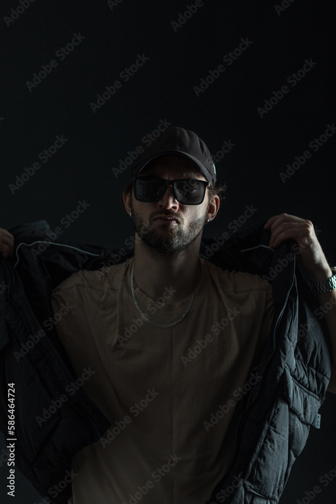 Fashionable handsome man with sunglasses and a cap in trendy clothes puts on a vest in the dark Fashionable handsome man with sunglasses and a cap in trendy clothes puts on a vest in the dark