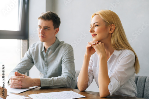 Portrait of Caucasian HR male and female managers interviewing unrecognizable male job seeker sitting at desk near window. Smiling young woman and man headhunters holding job interview with candidate.