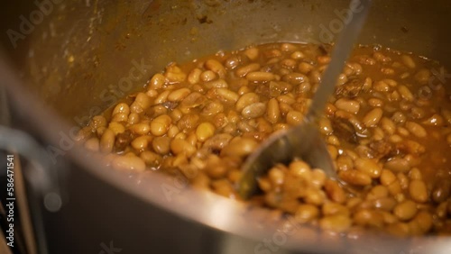 Large spoon stirs huge pot filled with barbecue pinto beans, slow motion close up 4K