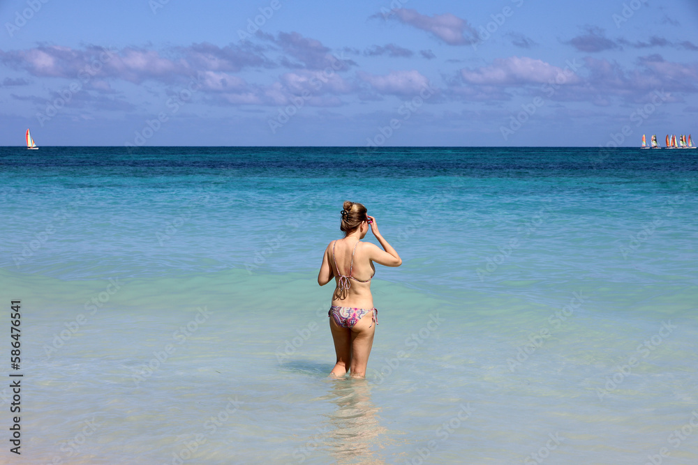 Woman in bikini standing blue sea water and looking to sailboats on horizon. Beach vacation on Caribbean islands