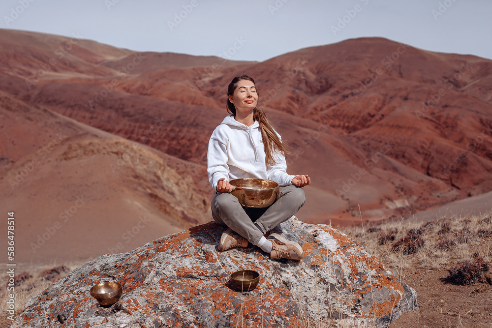 A smiling girl sits on a large stone and meditates in the lotus position with her eyes closed. At her feet lies a large musical bowl.