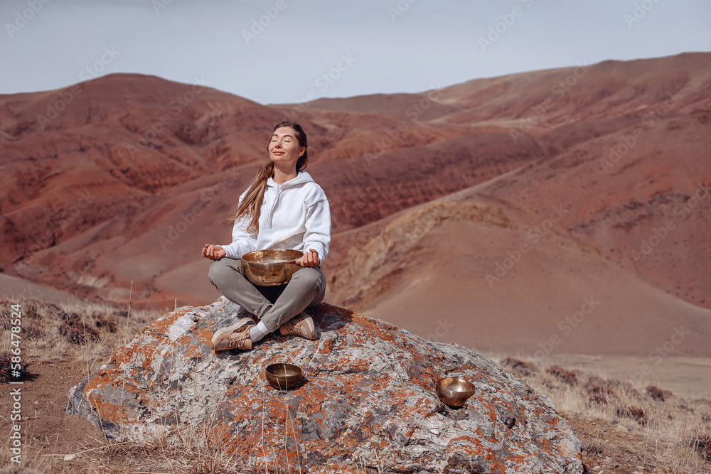 Girl sits on stone with singing bowl at her feet. Practice meditation in lotus position with closed eyes. Getting pleasure. Mountain background. Girl sits on stone with singing bowl at her feet. Practice meditation in lotus position with closed eyes. Getting pleasure. Mountain background.
