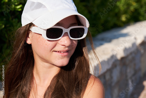 Teen girl with freckles in a white cap with sunglasses on the beach.