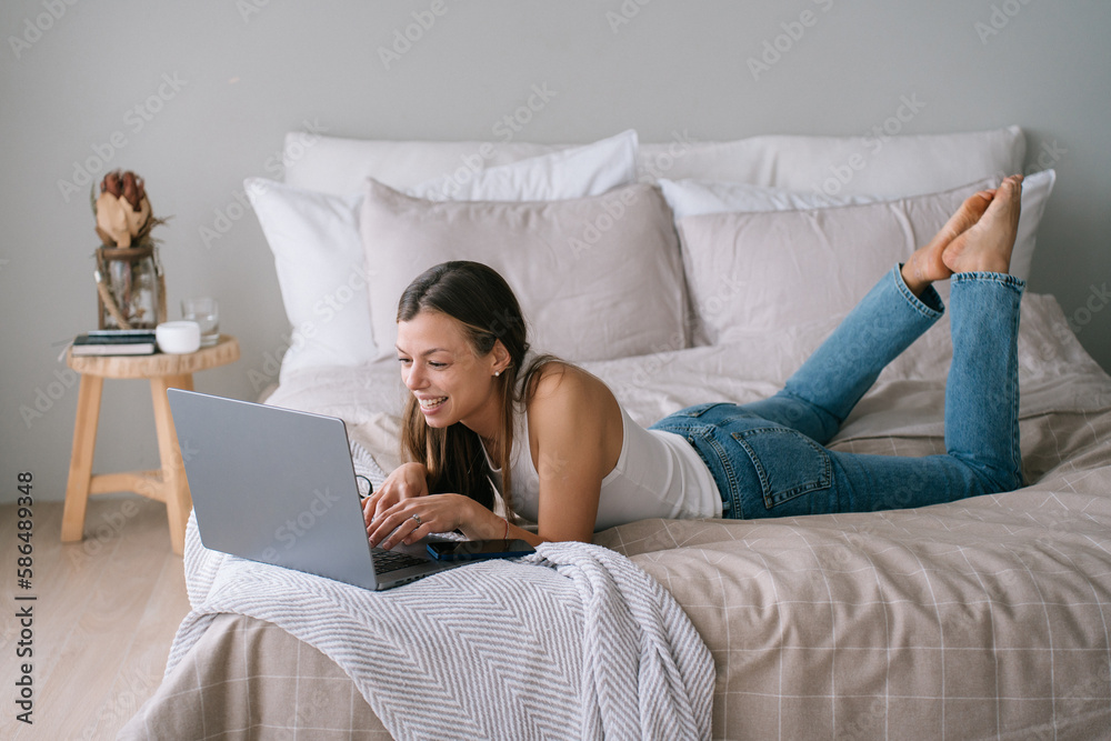 Adorable caucasian girl laying on bed with big cushions using laptop chatting with boyfriend by phone, makes video call toothy smiles. Happy hispanic young woman in blue jeans relaxing home.