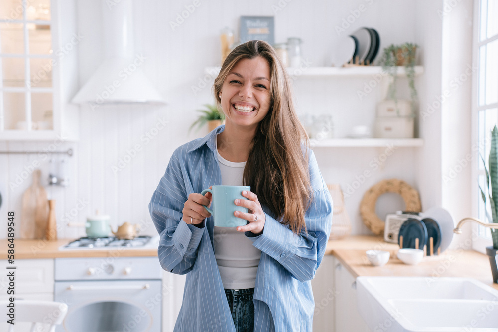 Happy caucasian female with long loose hair in jeans and blue shirt smiles holds cup of tea looks at camera happily enjoys weekend at cozy home. Pretty hispanic woman laughs against blurry kitchen Happy caucasian female with long loose hair in jeans and blue shirt smiles holds cup of tea looks at camera happily enjoys weekend at cozy home. Pretty hispanic woman laughs against blurry kitchen