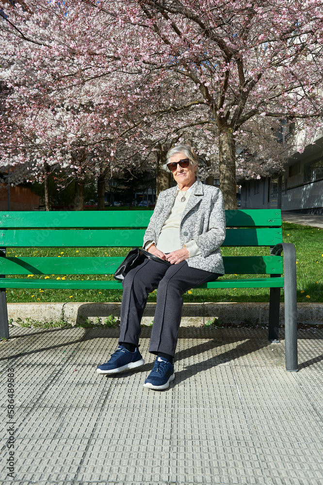 an eighty-year-old woman sunbathes in sunglasses sitting on a green wooden bench under a Japanese cherry tree