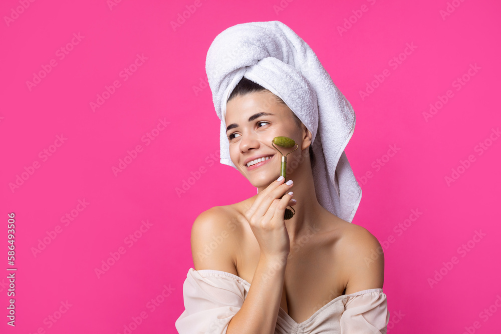 A beautiful cheerful attractive girl with a towel on her head holds a cosmetic roller near her face against a pink background. A beautiful cheerful attractive girl with a towel on her head holds a cosmetic roller near her face against a pink background.