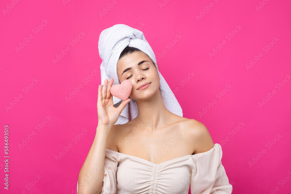 Beautiful cheerful attractive girl with a towel on her head, holds a sponge in the form of a pink heart.