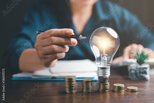 Businesswoman with lightbulb on coins and using calculator to calculate and money stack. Save energy and money with accounting finance in office concept. Idea of energy saving planning in home.