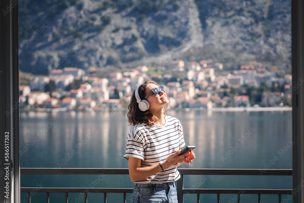Young happy woman listening to music using headphones while standing on the balcony with amazing lake and mountain views