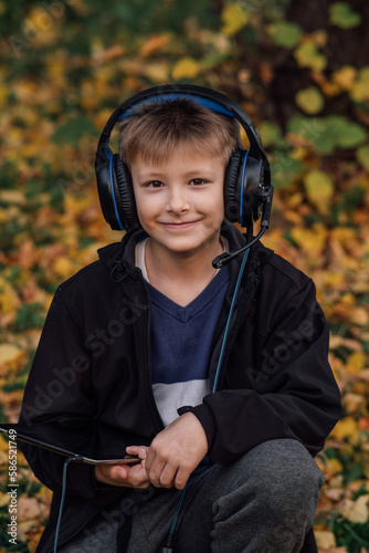 close-up portrait of a European blond boy in a black jacket with a tablet and headphones near a school, for online learning, a happy young man in a park. A happy schoolboy. Autumn photo, front view
