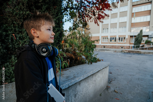 Portrait of European blond boy in black jacket with headphones looking at school, online learning, happy young man in school yard. Teenager in front of school, front view, open space. 