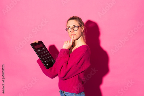 Thoughtful woman gesturing and holding calculator against pink background photo