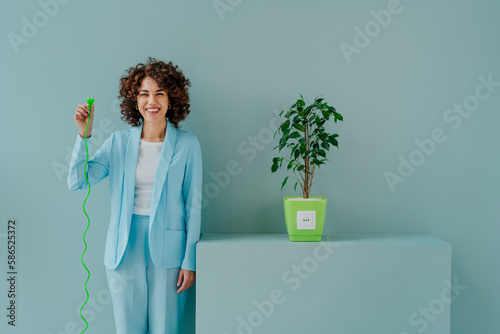 Happy woman with electric plug standing by potted plant in front of blue wall