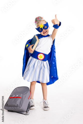Pre schooler daughte in super hero costume standing on white background holding book and grey backpack ready to school concept. © ABCreative