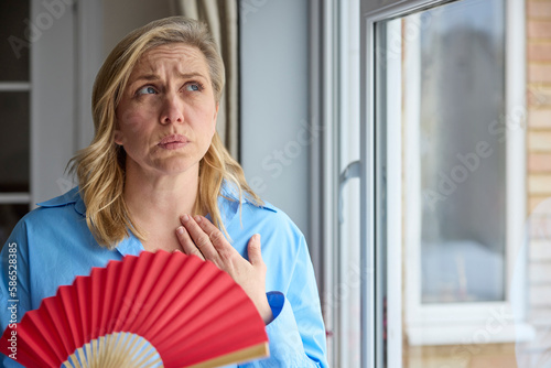 Menopausal Mature Woman Having Hot Flush At Home Cooling Herself With Handheld Paper Fan photo