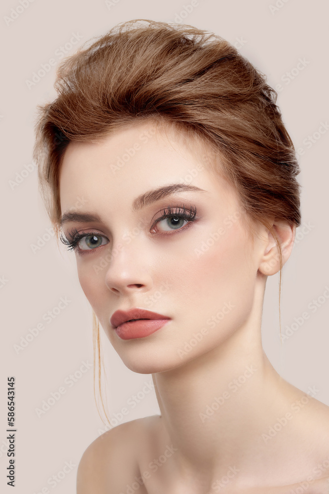Obraz premium Close-up shot of a young European woman with makeup and bouffant hairstyle. Beautiful girl with her hair pulled back is isolated on a beige background. Modern hairstyle on woman. Front view.