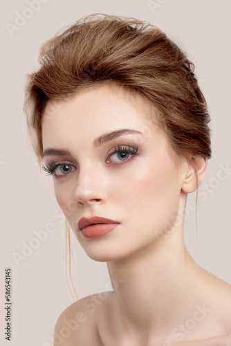 Close-up shot of a young European woman with makeup and bouffant hairstyle. Beautiful girl with her hair pulled back is isolated on a beige background. Modern hairstyle on woman. Front view.