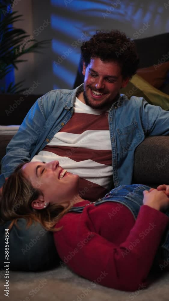Vertical view of young caucasian couple smiling and looking at each other, resting sitting on the carpet on the floor at home and leaning on the sofa on a romantic date Husband and wife relaxing. High Vertical view of young caucasian couple smiling and looking at each other, resting sitting on the carpet on the floor at home and leaning on the sofa on a romantic date Husband and wife relaxing. High