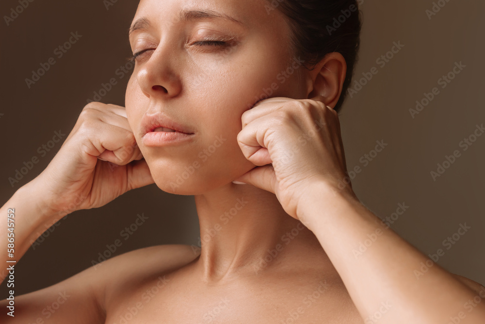 Cropped shot of young caucasian woman touching face with hands massaging her jaw isolated on a dark brown background. Rejuvenation, facelift, facefitness, skin care. Cosmetology and beauty concept Cropped shot of young caucasian woman touching face with hands massaging her jaw isolated on a dark brown background. Rejuvenation, facelift, facefitness, skin care. Cosmetology and beauty concept