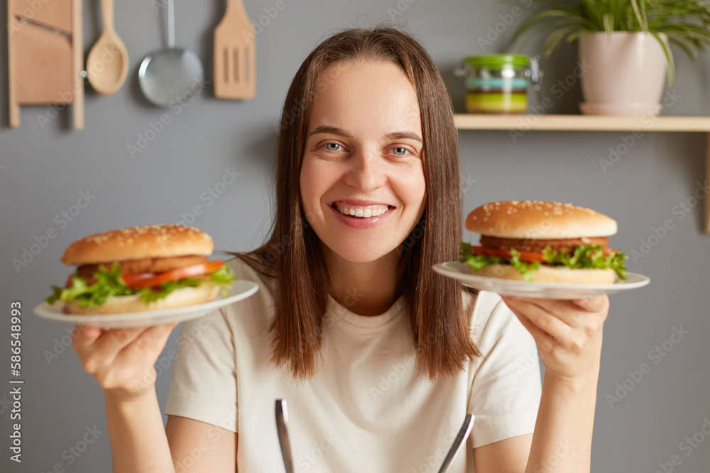 Indoor shot of smiling cheerful Caucasian woman holding two delicious fresh burgers, posing in kitchen, breaking diet, feels extremely hungry, looking at camera with happy face.
