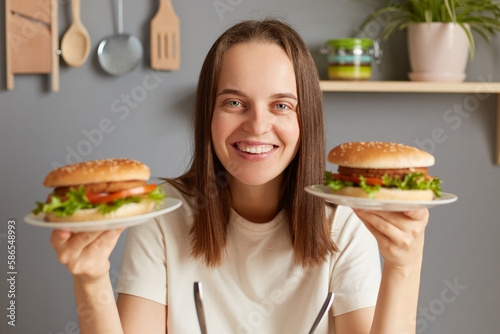 Indoor shot of smiling cheerful Caucasian woman holding two delicious fresh burgers  posing in kitchen  breaking diet  feels extremely hungry  looking at camera with happy face.