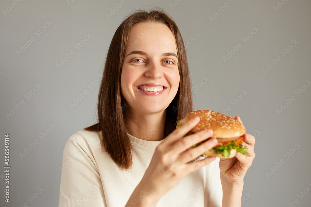 Portrait of smiling attractive woman holding delicious fresh burger, eating junk food snack, looks at camera with overjoyed expression, having cheat meal, enjoying tasty sandwich.