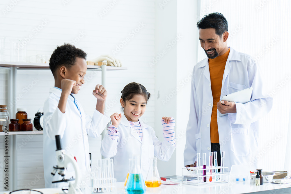 .In chemistry classroom with many laboratory tools on shelves and table. A male teacher in white lab coat confirms experimental result from a young African boy and little Asian girl with joy together. .In chemistry classroom with many laboratory tools on shelves and table. A male teacher in white lab coat confirms experimental result from a young African boy and little Asian girl with joy together.